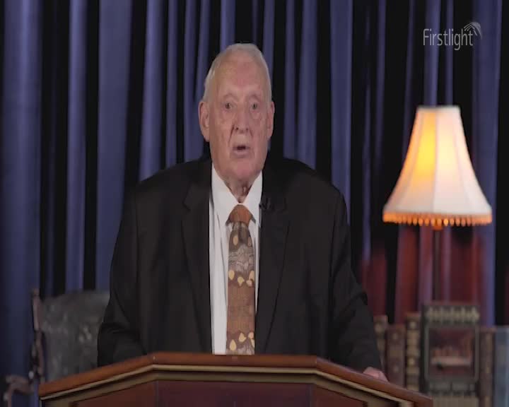 An older man in a dark suit stands behind a wooden podium, speaking. The Firstlight logo sits in the upper right corner, and a lit lamp glows beside him.
