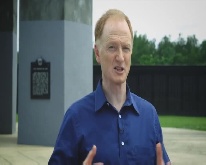 A man in a blue shirt is gesturing with his hands, speaking directly to the camera. Behind him, there's a dark fence and a stone structure with a plaque, likely in New Zealand.
