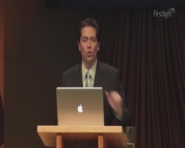 A man in a suit stands at a podium, gesturing with his right hand. A laptop sits in front of him, the Apple logo glowing.
