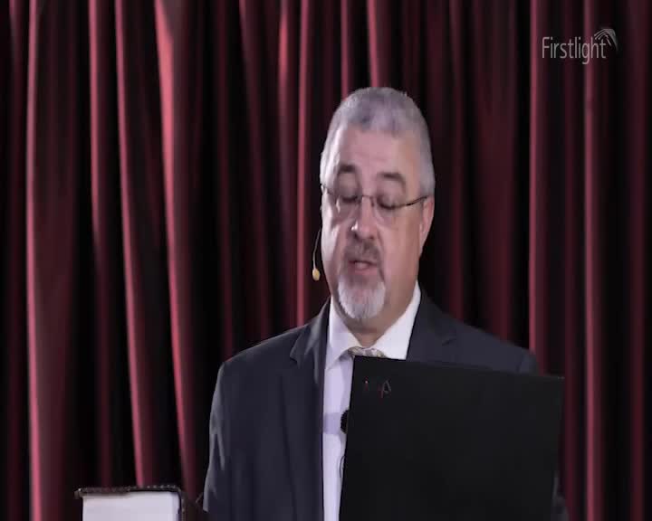A man in a suit, wearing glasses and a lavalier microphone, is speaking, possibly reading from a laptop. Behind him, a burgundy curtain provides a backdrop for what appears to be a presentation from Firstlight.
