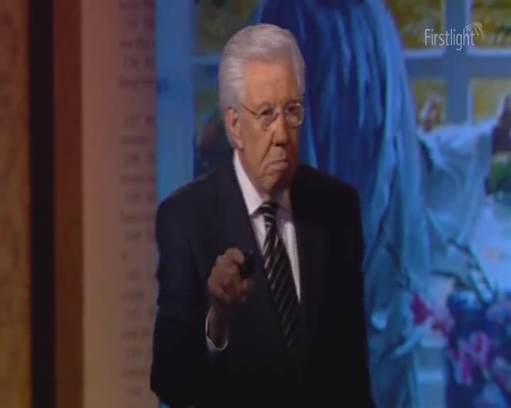 The man in the suit, with his silver hair and glasses, holds a small black object and points directly at the camera. Behind him, a blurred image of a floral arrangement and a figure in a white robe is visible on the screen, with the "Firstlight" logo in the upper right corner.
