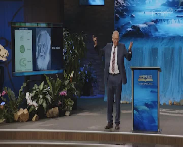 A man in a suit stands center stage, gesturing with both hands as he speaks. Behind him, a screen displays a portrait and diagrams, while a backdrop shows a waterfall, and the Firstlight logo.
