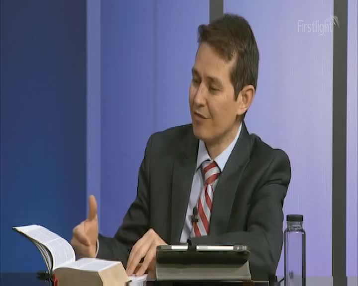 The man in the suit gestures with his right hand as he speaks, a large book open on the desk in front of him. A Firstlight logo is visible in the background.
