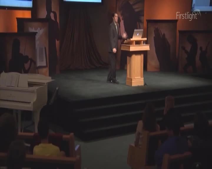A man in a suit stands at a wooden podium, a laptop perched on top. He's facing the audience, likely about to speak in front of a Firstlight backdrop.
