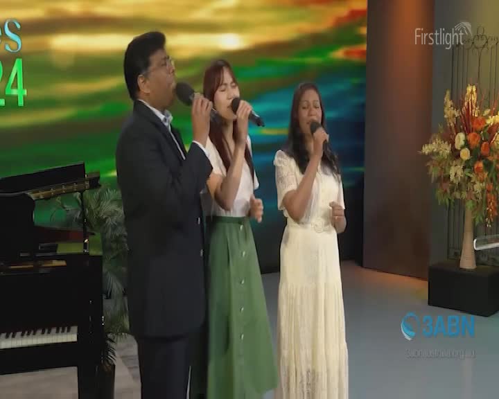 Three people stand close together, each holding a microphone and singing on a stage. A man in a dark suit and two women in light-colored dresses are the focus, with a grand piano to the left and a floral arrangement to the right.
