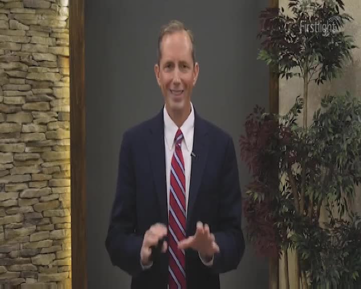 A man in a suit stands in front of a stone wall, gesturing with his hands. The Firstlight logo is visible in the top right corner.
