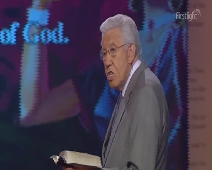 The man in the gray suit is speaking, his mouth open as he holds a book. A "Firstlight" logo is visible in the corner, and a backdrop with text is behind him.
