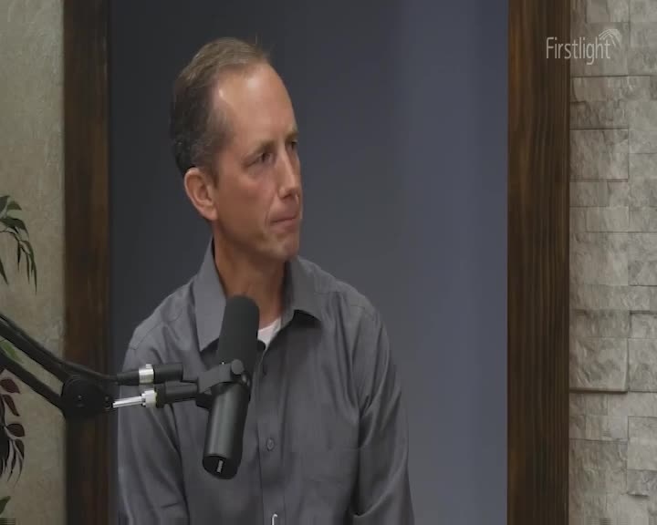 A man in a grey shirt sits, looking slightly off-camera, a microphone positioned in front of him. The Firstlight logo is visible in the upper right corner, suggesting a broadcast setting.
