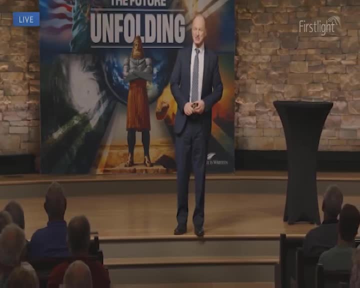 A man in a suit stands on a stage, speaking to an audience. Behind him, a large banner with the words "The Future Unfolding" is displayed, and the event is being broadcast "LIVE" by Firstlight.
