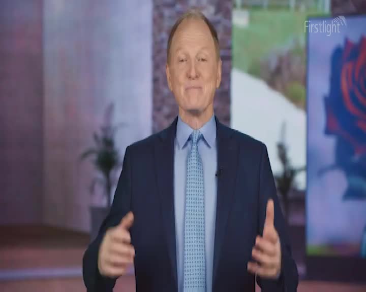 The man in the suit gestures with both hands, his expression friendly. Behind him, a Firstlight logo is visible, along with a backdrop of a rose and a landscape.
