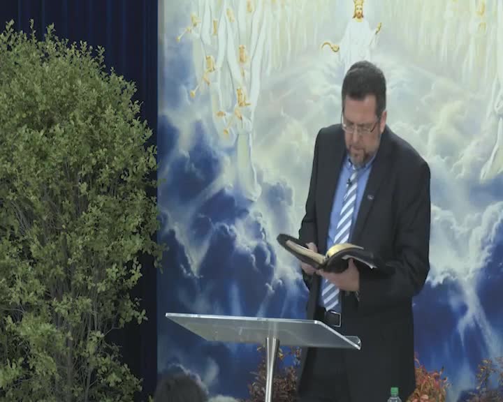 A man in a suit stands at a lectern, intently reading from a book with gold-edged pages. Behind him, a painted backdrop depicts figures in white robes against a cloudy sky.
