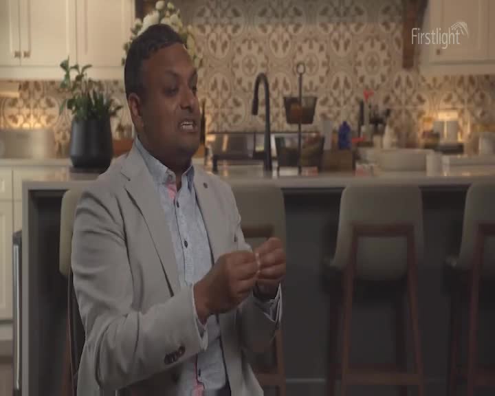 A man in a light grey blazer gestures with his hands, speaking animatedly. Behind him, a kitchen island with bar stools and a patterned backsplash provides the backdrop.
