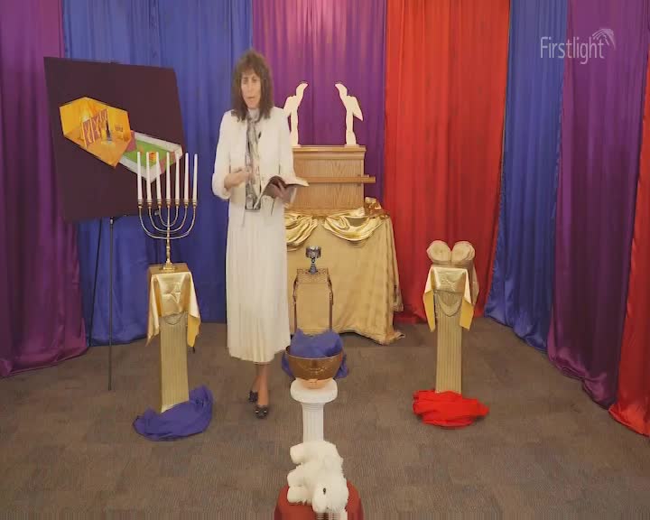 A woman in a white dress stands center stage, holding a book and speaking. Behind her, a backdrop of colorful drapes and various symbolic objects fill the space.
