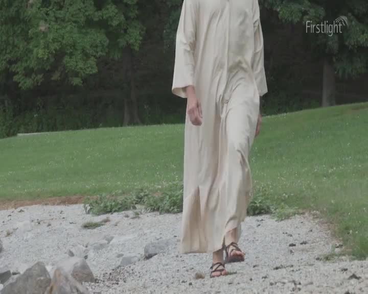 A person in a long, cream-colored robe walks toward me on a gravel path. Their sandals make a soft sound as they step, the fabric of the robe swaying with each stride.
