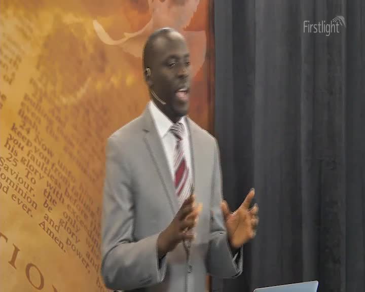 A man in a gray suit gestures with his hands as he speaks, a microphone clipped to his lapel. Behind him, a backdrop features scriptural text and the "Firstlight" logo.
