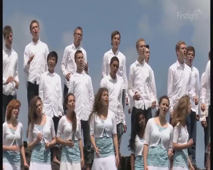 A choir, dressed in white, is singing outdoors under a bright sky. Their voices rise and fall as they stand on a rocky outcrop, the Firstlight logo visible in the corner.
