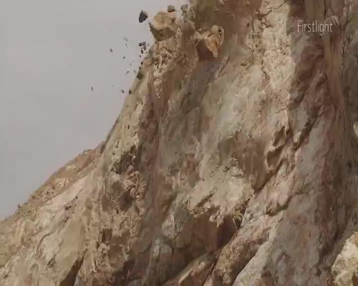 Chunks of rock are tumbling down the cliff face, some already broken into smaller pieces. Dust is rising as the sun shines on the rock's surface.
