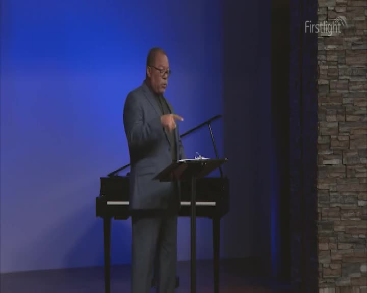 A man in a dark suit gestures emphatically from behind a lectern, his words echoing in the Firstlight studio. Beside him, a grand piano sits silently against a backdrop of deep blue.
