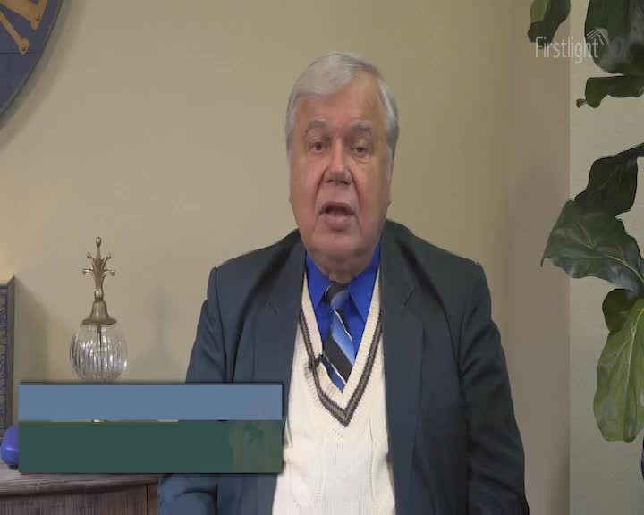 A man in a suit and sweater vest is speaking, his mouth moving as he addresses the camera. The Firstlight logo is visible in the background, along with a potted plant.
