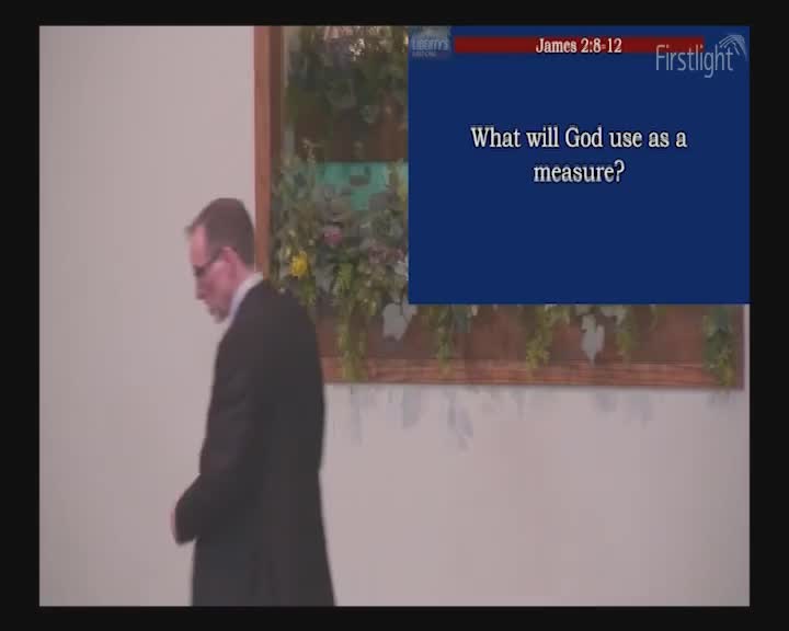 A man in a suit walks past, his head bowed. A blue screen with text from Firstlight hangs on the wall beside him.
