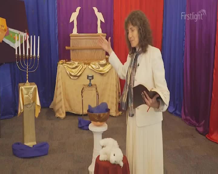 A woman in a cream suit gestures towards a table draped in gold fabric, a small bible in her other hand. Behind her, a set is decorated with religious props, including a menorah and a model of the Ark of the Covenant.
