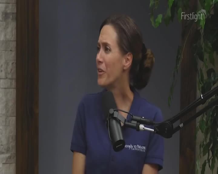 A woman in a dark blue shirt is speaking, her expression animated. A microphone is positioned in front of her, and the word "Firstlight" is visible in the upper right corner.
