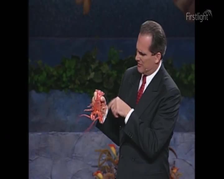 A man in a suit gestures towards a bright red, spiky crayfish he holds in his hand. The Firstlight logo is visible in the corner.
