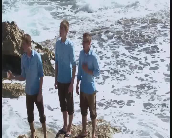 Three men in matching blue shirts and brown shorts stand on a rocky shore, facing the camera. The ocean churns behind them, its foamy white crests contrasting with the darker rocks.
