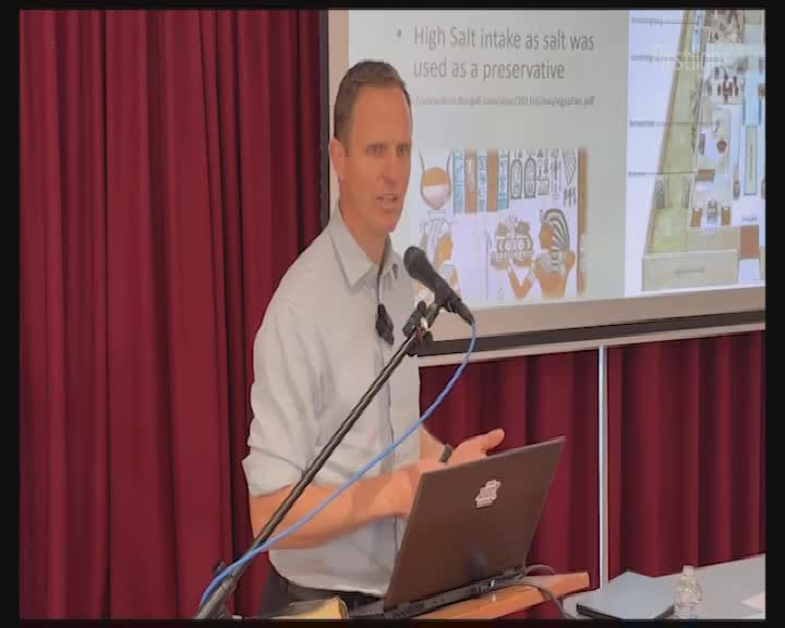 A man in a light blue shirt gestures with his hands, speaking into a microphone while standing behind a laptop. Behind him, a projector screen displays text and images, likely part of a presentation.
