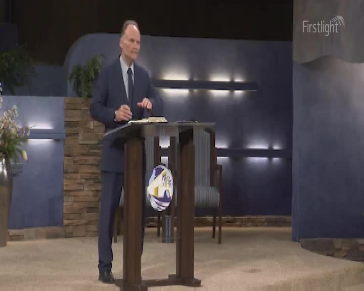 A man in a suit stands behind a podium, gesturing with his hands as he speaks. The Firstlight logo is visible in the corner of the room.
