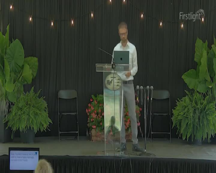 A man in a light shirt and grey trousers stands at a lectern, looking at a laptop. The stage is decorated with lush green plants and a string of small lights overhead.
