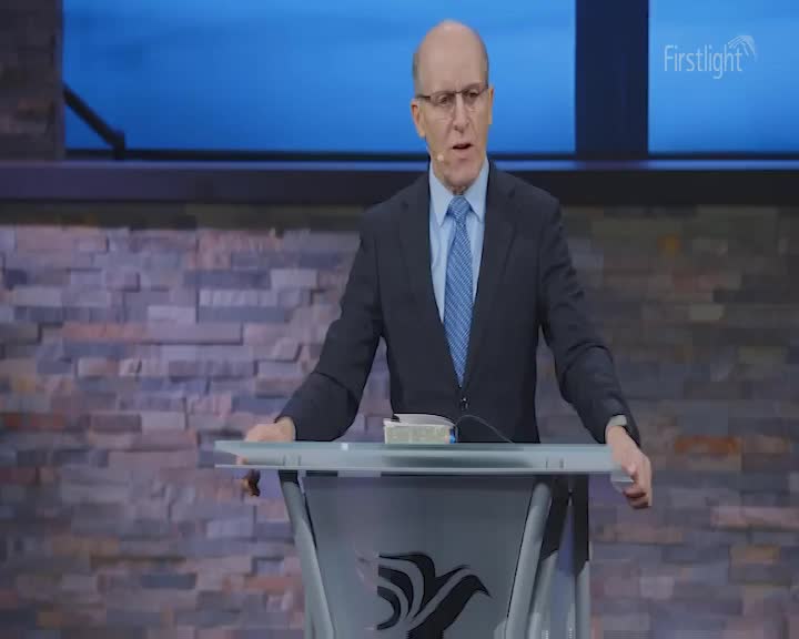 A man in a suit stands behind a podium, his hands resting on its surface. He speaks with conviction, his gaze directed forward.