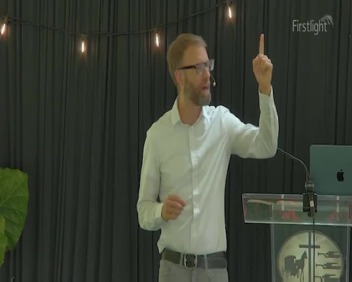 A man in a white shirt gestures with his right hand, index finger pointing upwards. He stands before a dark curtain, a laptop on a podium beside him.
