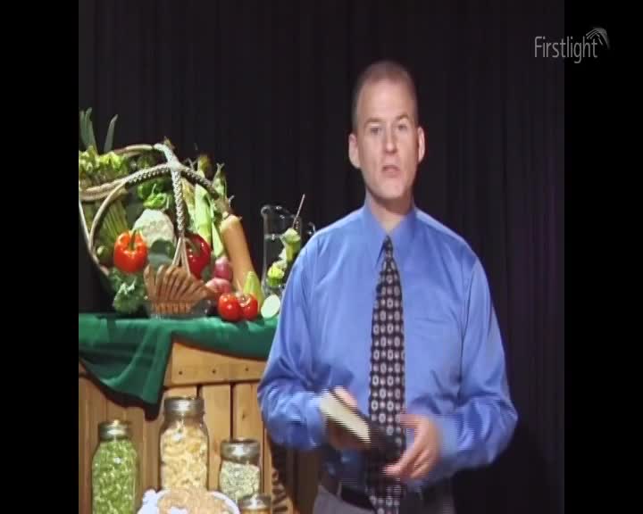A man in a blue shirt and patterned tie stands before a display of fresh produce. He holds a book, his gaze directed forward.