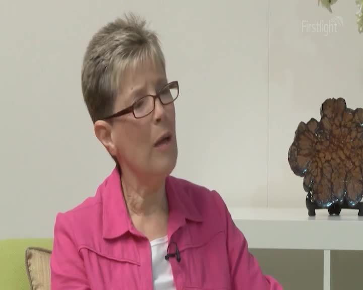 A woman with short, grey hair and glasses speaks, her pink jacket open over a white top. Beside her, a decorative piece with a rough, textured surface sits on a shelf.