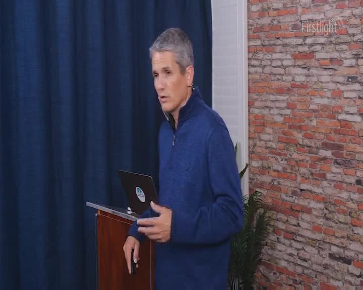 A man in a blue zip-up shirt speaks with his hands, gesturing towards a laptop on a podium. The backdrop is a dark blue curtain and a brick wall.