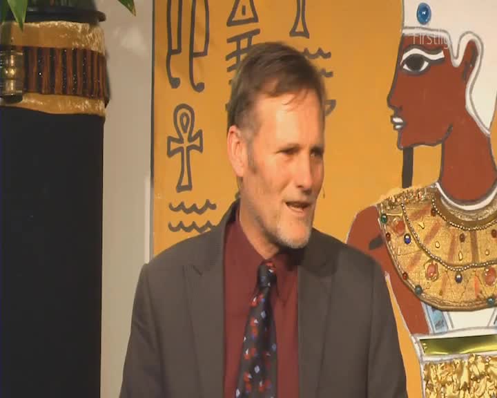 The man in the suit speaks, his voice carrying over the gentle hum of the early morning. Behind him, a stylized Egyptian figure stands guard, bathed in the soft glow of firstlight.