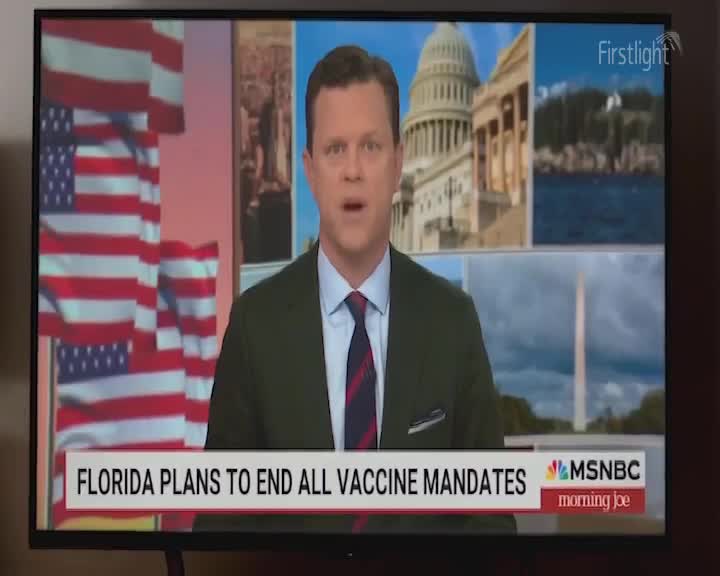 The presenter speaks with a backdrop of American flags and government buildings. A chyron at the bottom reads "FLORIDA PLANS TO END ALL VACCINE MANDATES."
