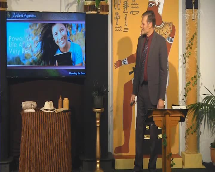 A man in a suit gestures towards a large mural of an ancient Egyptian figure. On a screen above, a young woman smiles broadly, holding a book.
