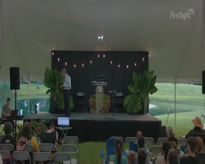 A man in a light blue shirt stands on a stage, gesturing with his hands. The Firstlight logo appears in the upper right corner.