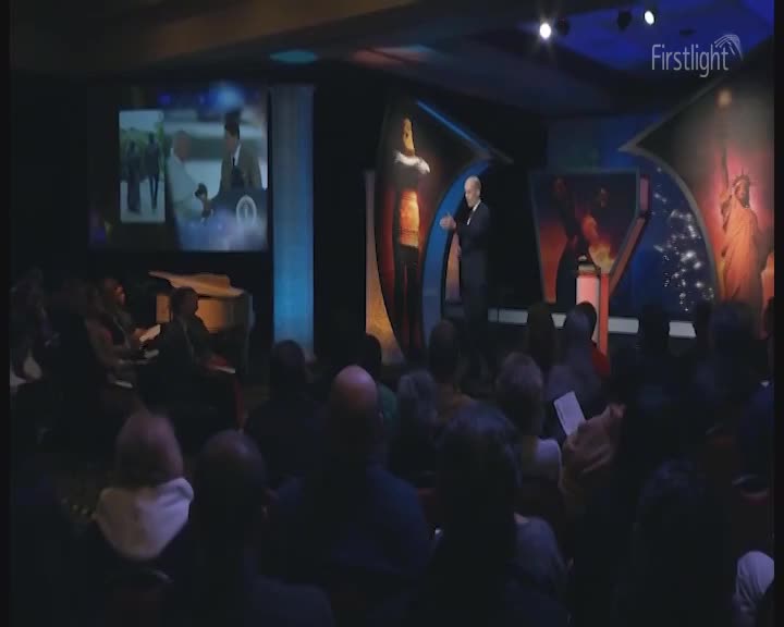 A man in a suit stands on a stage, gesturing as a video plays on a screen behind him. The audience sits in the dim light, some holding papers. A man in a suit stands on a stage, gesturing as a video plays on a screen behind him. The audience sits in the dim light, some holding papers.