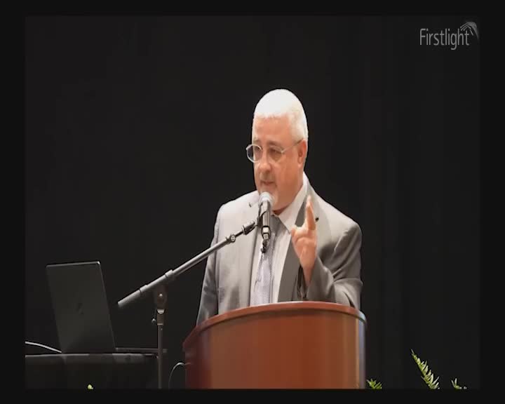 A man in a suit stands at a podium, gesturing with his right hand as he speaks into a microphone. A laptop sits open to his left, and a sprig of greenery is visible near the bottom right. A man in a suit stands at a podium, gesturing with his right hand as he speaks into a microphone. A laptop sits open to his left, and a sprig of greenery is visible near the bottom right.