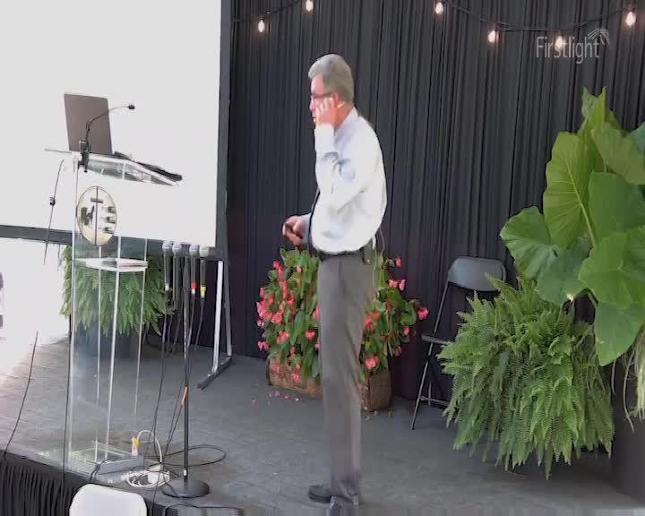 A man in a light blue shirt and grey trousers stands by a podium, holding his phone to his ear. Behind him, a dark curtain is strung with fairy lights, and lush green plants spill from pots. A man in a light blue shirt and grey trousers stands by a podium, holding his phone to his ear. Behind him, a dark curtain is strung with fairy lights, and lush green plants spill from pots.