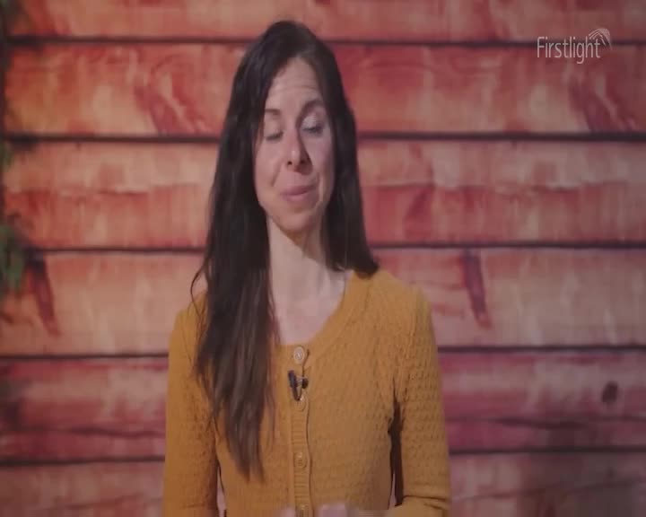 A woman with long, dark hair stands before a wooden backdrop. She wears a mustard-yellow cardigan. A woman with long, dark hair stands before a wooden backdrop. She wears a mustard-yellow cardigan.
