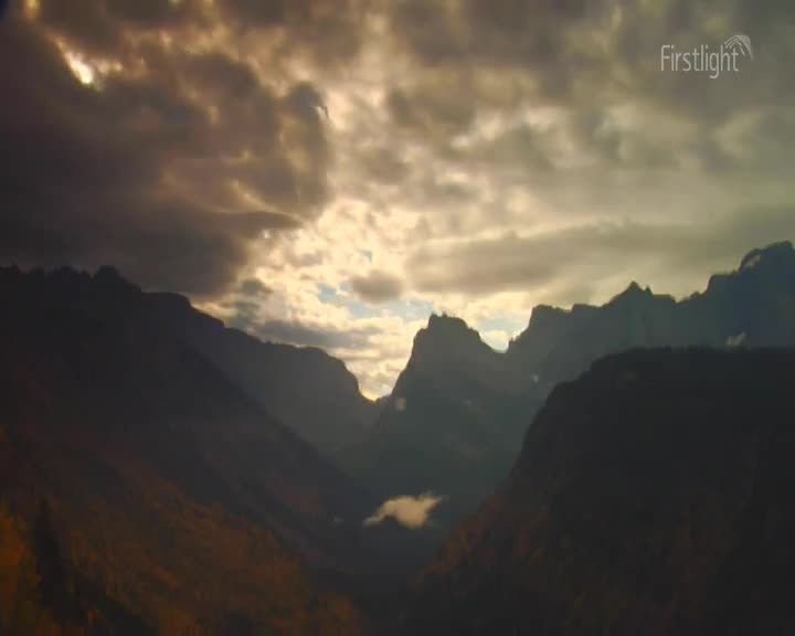 The sun breaks through heavy clouds, illuminating jagged peaks in the distance. A valley floor, tinged with autumn colours, slopes away into shadow. The sun breaks through heavy clouds, illuminating jagged peaks in the distance. A valley floor, tinged with autumn colours, slopes away into shadow.