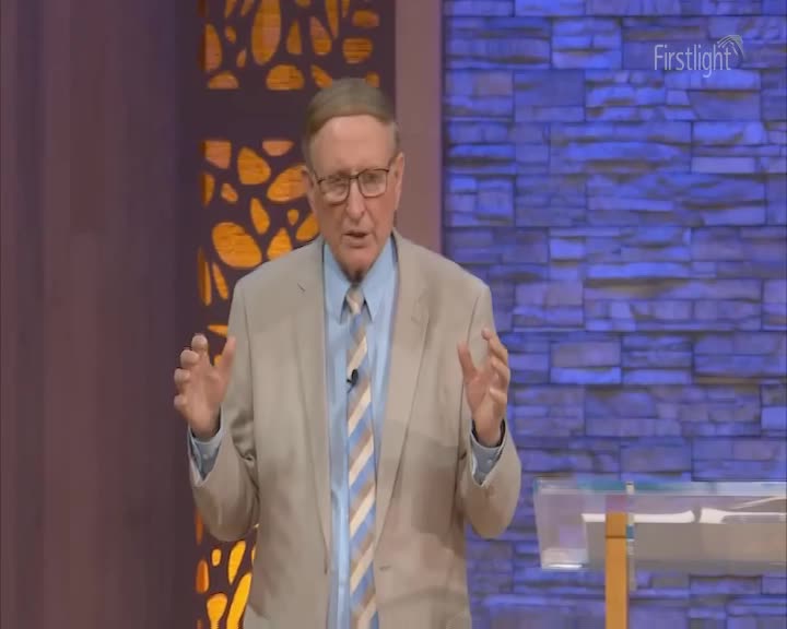 A man in a light suit and striped tie gestures with both hands as he speaks. He stands before a backdrop of textured blue bricks, with a patterned screen to his left.