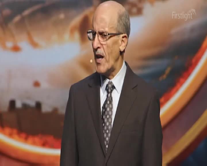 A man in a dark suit and patterned tie speaks animatedly, his mouth open. Behind him, a large screen displays a fiery, abstract landscape, hinting at the early morning light of New Zealand.