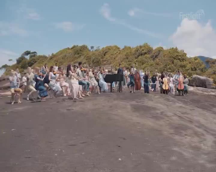 A string orchestra plays on a rocky New Zealand coastline as the sun begins to rise. Cellos and violins are held aloft, their bows moving in unison against the backdrop of lush green hills.