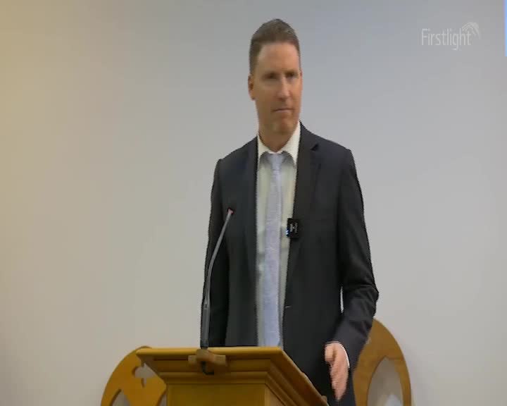 A man in a dark suit and light blue tie stands behind a wooden lectern. He gestures with his right hand as he speaks, his expression serious.