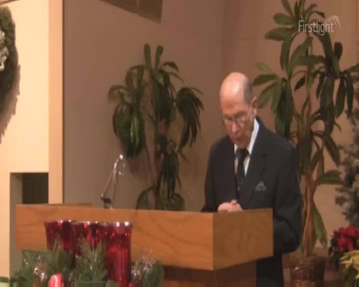 A man in a suit stands at a wooden lectern, his hands clasped. Lush green plants frame the scene, and a small Christmas tree with red poinsettias sits to the right.